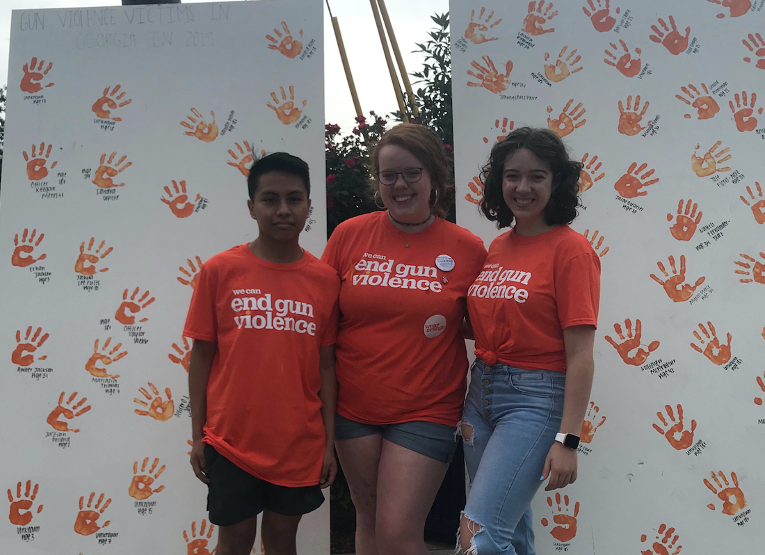 Image from the Wear Orange art project,
      students smiling in front of two white canvasses with orange handprints and names.